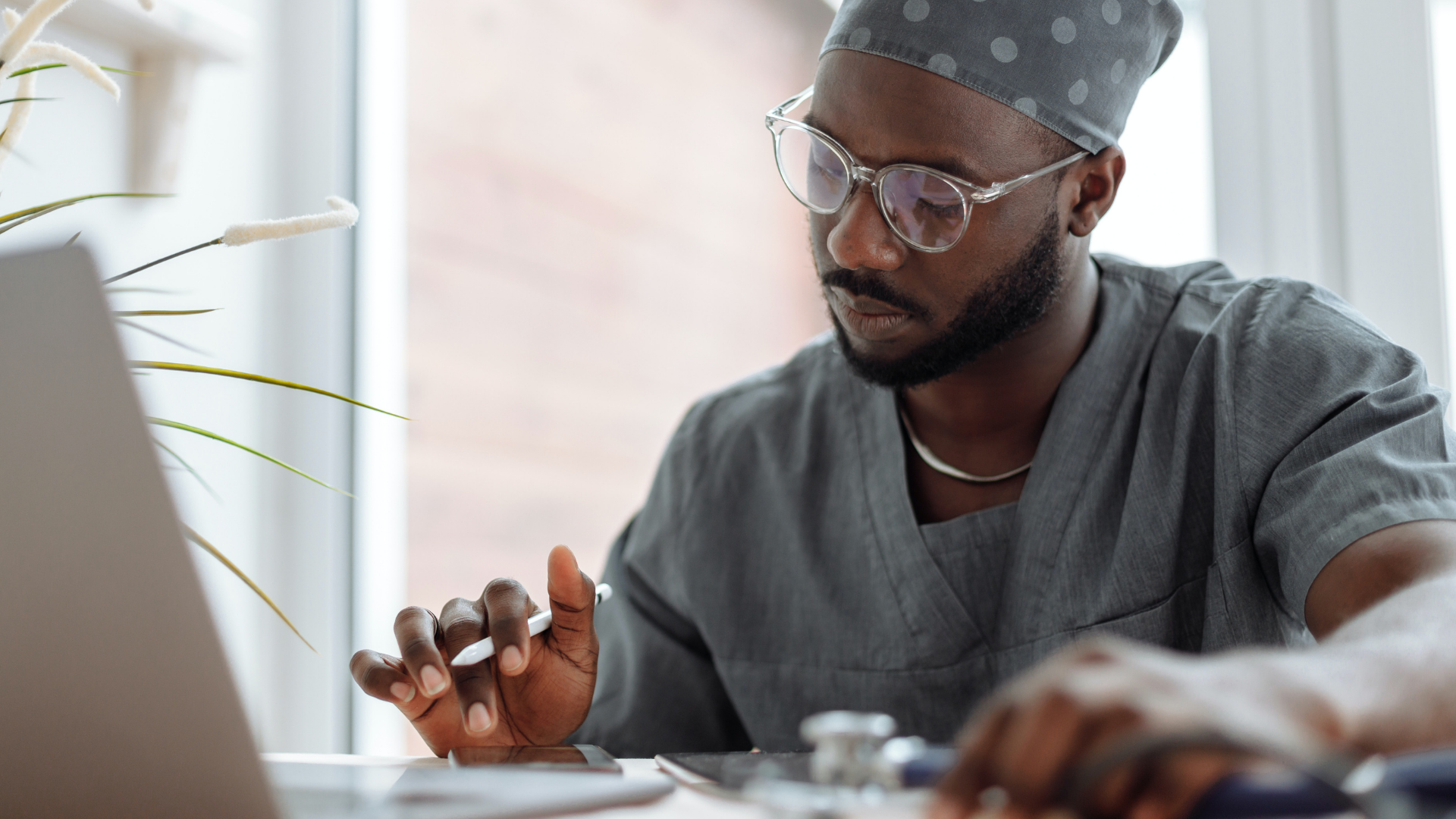 Doctor wearing grey scrubs and glasses while working on his laptop